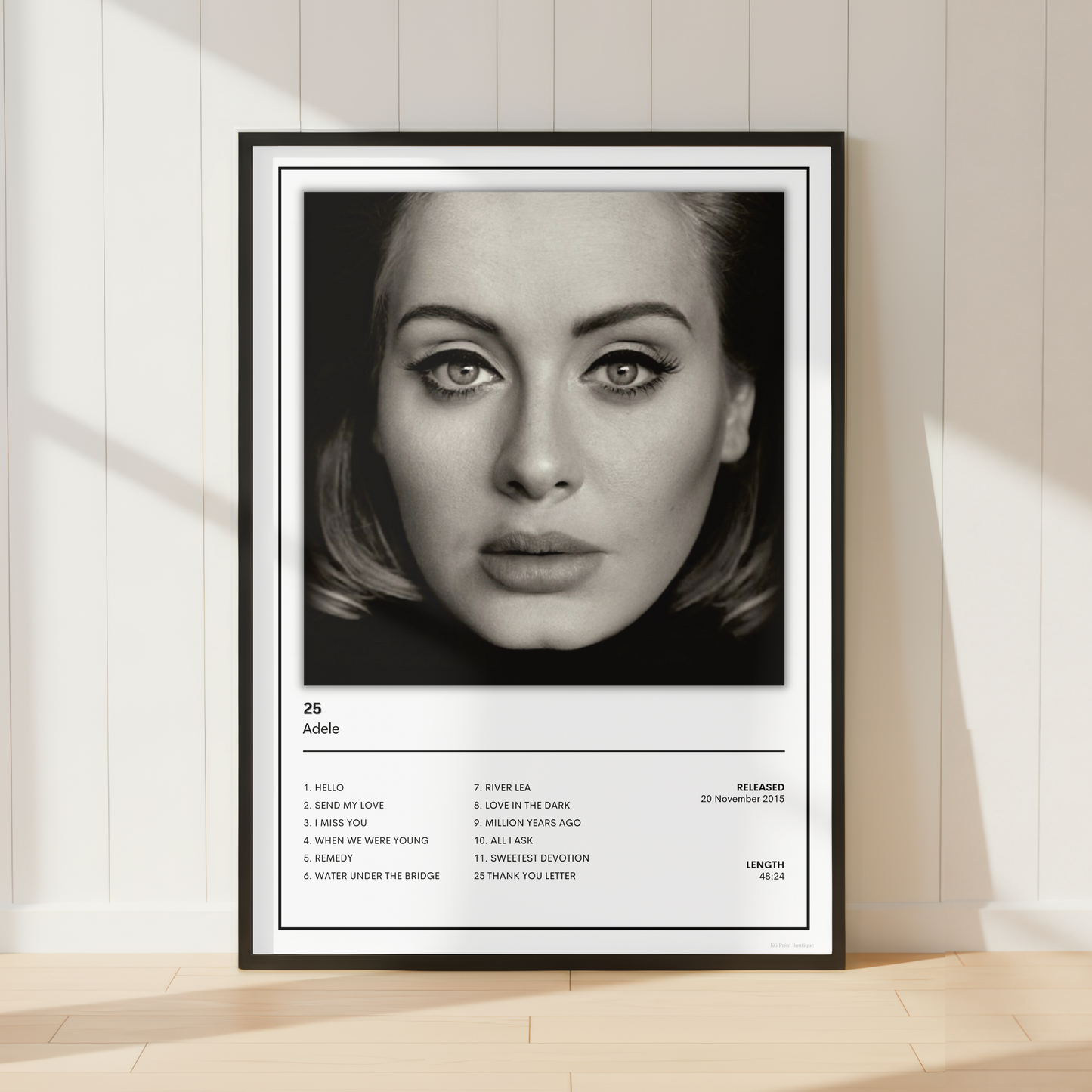 A framed black and white music poster of Adele's album '25' leaning against a cream-colored paneled wall on a light wood floor.
