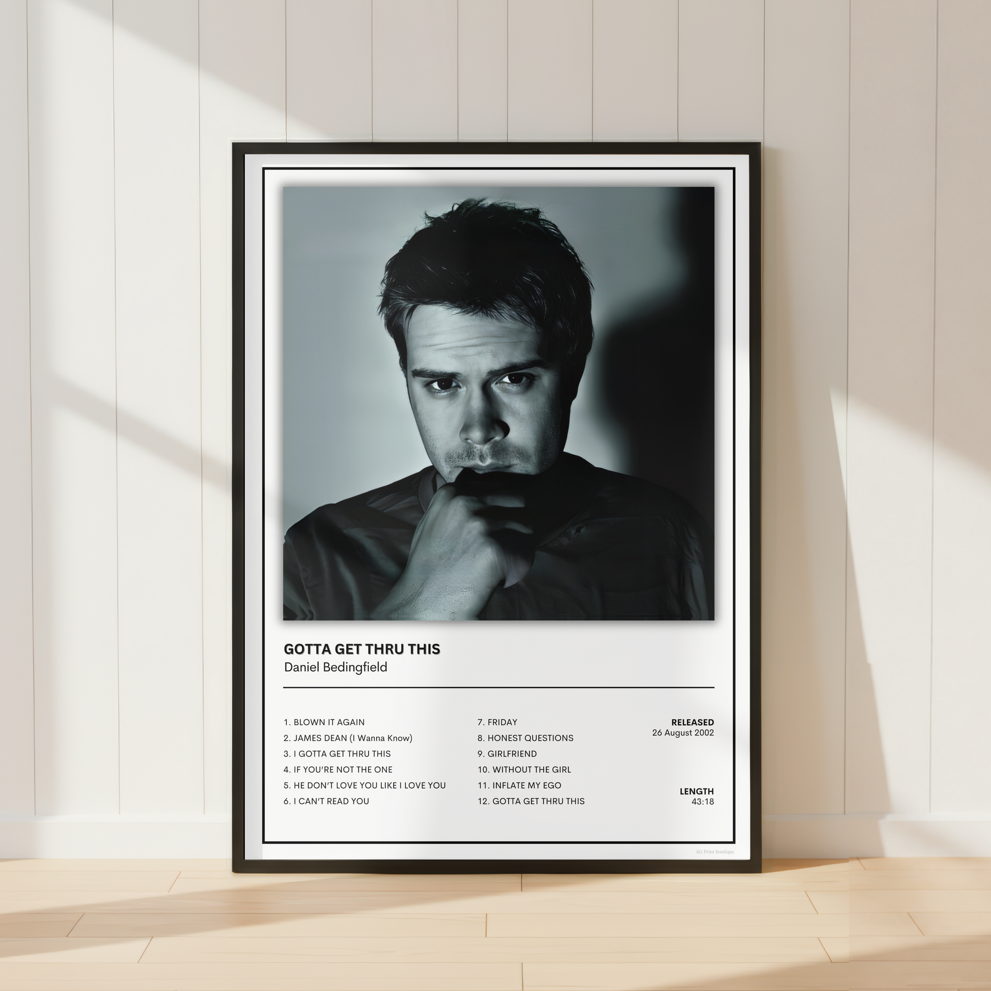 A framed black and white music poster of Daniel Bedingfield's song 'Gotta Get Thru This' leaning against a white paneled wall on a light wood floor.