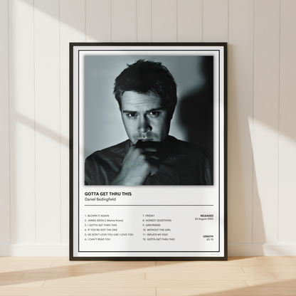 A framed black and white music poster of Daniel Bedingfield's song 'Gotta Get Thru This' leaning against a white paneled wall on a light wood floor.