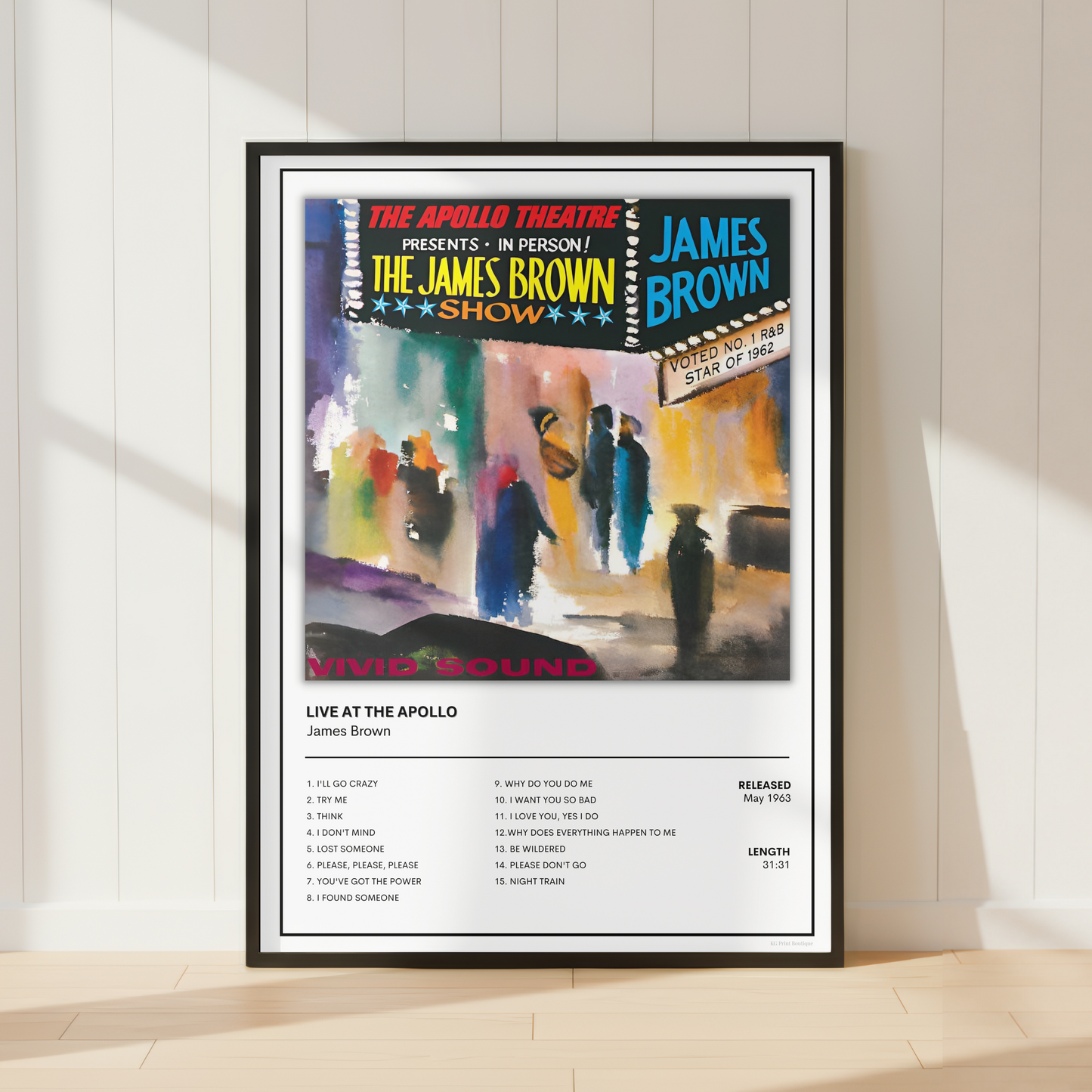 A framed concert poster for James Brown's 'Live at the Apollo' is displayed leaning against a white paneled wall on a light wood floor. The poster features a colorful painted performance scene with audience members and a black-framed tracklist section below.