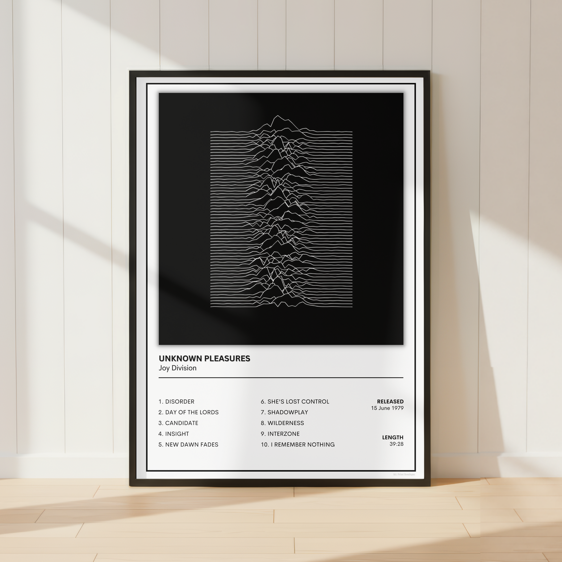 A framed music poster for the band Joy Division, featuring their 'Unknown Pleasures' album artwork, displayed on a light wood floor against a white paneled wall with soft window shadows.