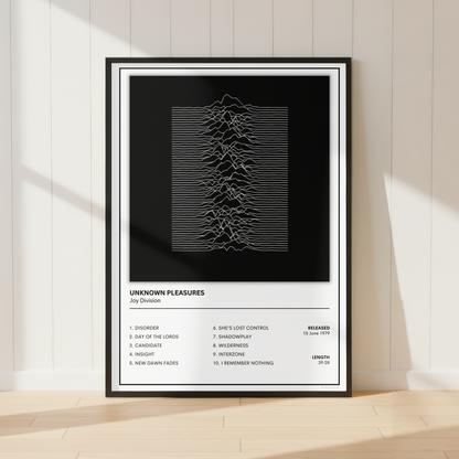 A framed music poster for the band Joy Division, featuring their 'Unknown Pleasures' album artwork, displayed on a light wood floor against a white paneled wall with soft window shadows.