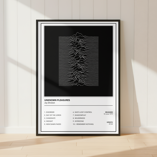 A framed music poster for the band Joy Division, featuring their 'Unknown Pleasures' album artwork, displayed on a light wood floor against a white paneled wall with soft window shadows.