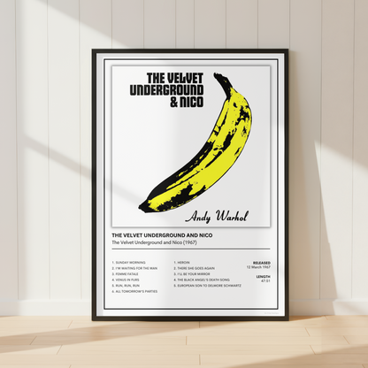 A framed music poster of The Velvet Underground & Nico featuring a large yellow banana graphic and tracklist, leaning against a white paneled wall on a light wood floor.