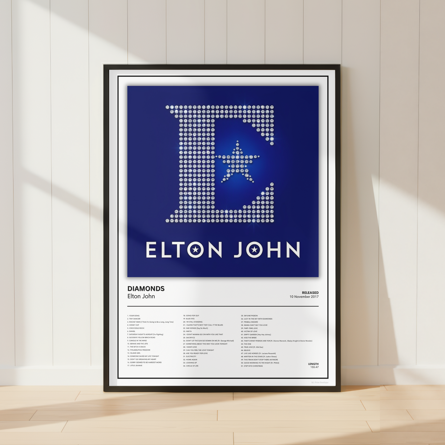 A framed music poster of Elton John's album 'Diamonds' leaning against a white paneled wall on a light wood floor. The poster features a large rhinestone-style letter 'E' with a star on a dark blue background, with 'ELTON JOHN' in white text below, and the tracklist on a white background below.