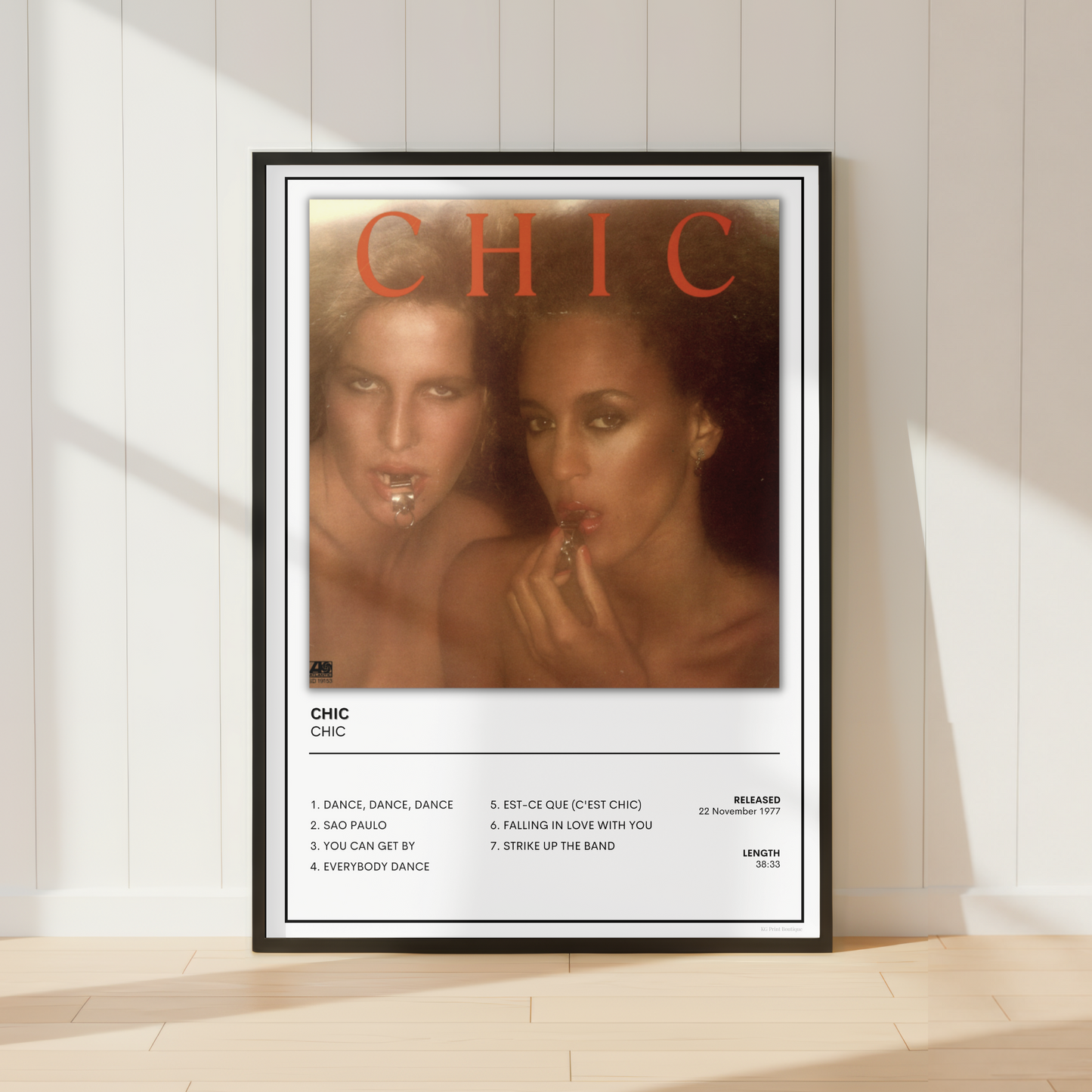 A framed music poster of the band CHIC, styled leaning against a white paneled wall on a light wood floor with soft window shadows.
