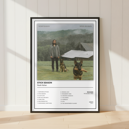 A framed poster of Noah Kahan's 'Stitck Season', featuring a tracklist and album art, leaning against a white paneled wall on a light wood floor.