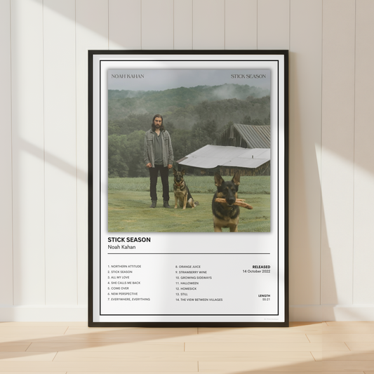 A framed poster of Noah Kahan's 'Stitck Season', featuring a tracklist and album art, leaning against a white paneled wall on a light wood floor.
