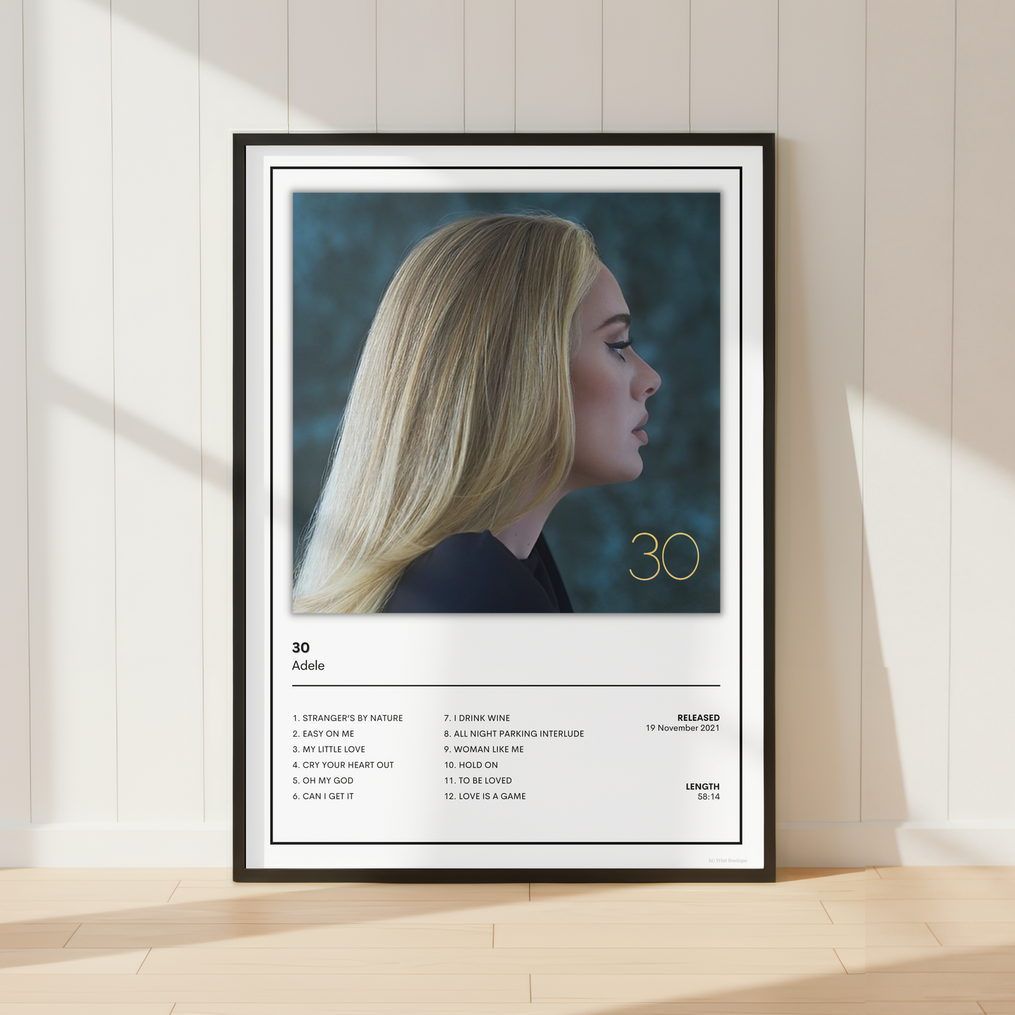 A framed vertical photo print leaning against a white paneled wall on a light wood floor. The print shows a blonde woman in profile with the number 30 and a small tracklist design below, in a black frame.