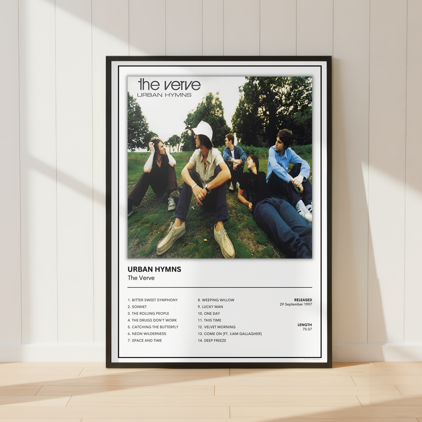A framed music poster for 'The Verve - Urban Hymns' leaning against a white paneled wall on a light wood floor, with soft sunlight casting shadows.