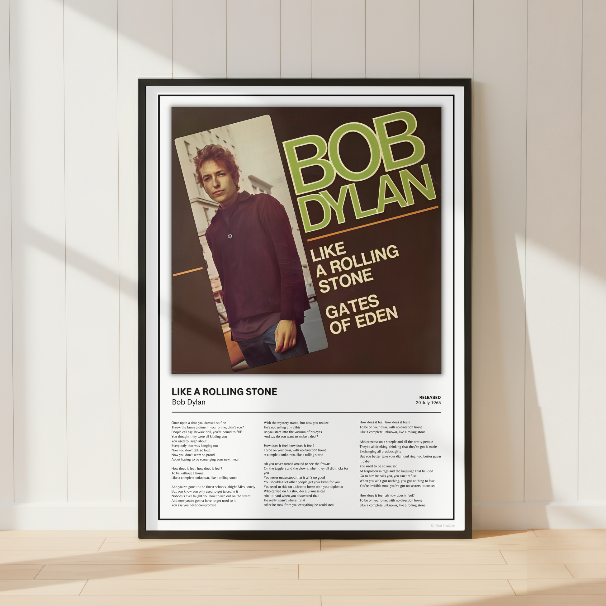 A framed Bob Dylan 'Like a Rolling Stone' poster leaning against a white paneled wall on a light wood floor, with soft window shadows cast across the scene.