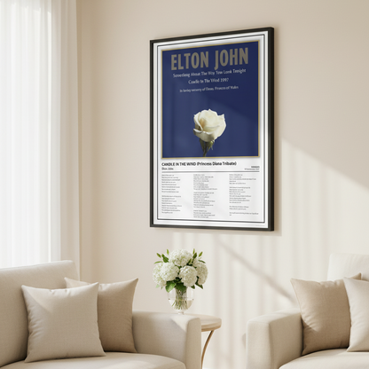 A framed Elton John tribute poster leaning against a white paneled wall on a light wood floor. The poster features a dark blue background with the text 'ELTON JOHN' and a single white rose, plus song lyrics at the bottom.