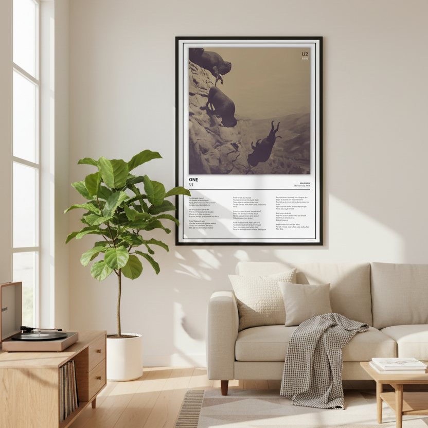 A framed art print leaning against a white paneled wall on a light wood floor. The print features a sepia-toned photograph of two figures on a cliff, with text below titled 'ONE' and additional smaller text.