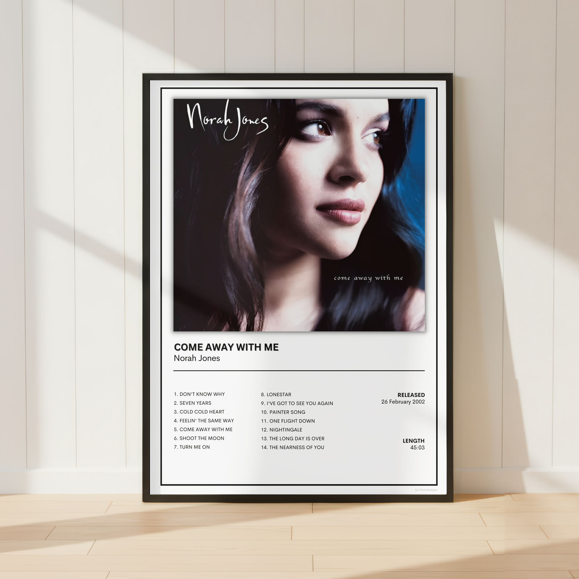 A framed music poster of Norah Jones' album 'Come Away With Me' leaning against a white paneled wall on a light wood floor, with soft sunlight casting shadows.
