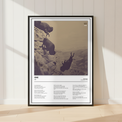 A framed art print leaning against a white paneled wall on a light wood floor. The print features a sepia-toned photograph of two figures on a cliff, with text below titled 'ONE' and additional smaller text.