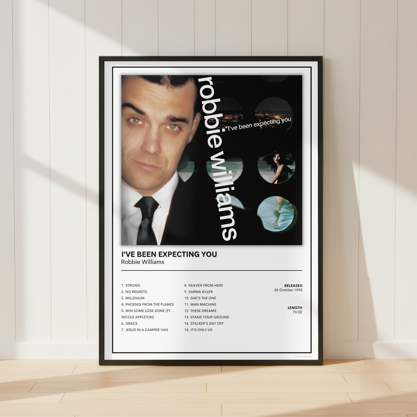 A framed poster of Robbie Williams leaning against a white paneled wall on a light wood floor, with soft diagonal shadows across the scene.