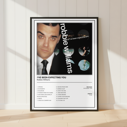 A framed poster of Robbie Williams leaning against a white paneled wall on a light wood floor, with soft diagonal shadows across the scene.