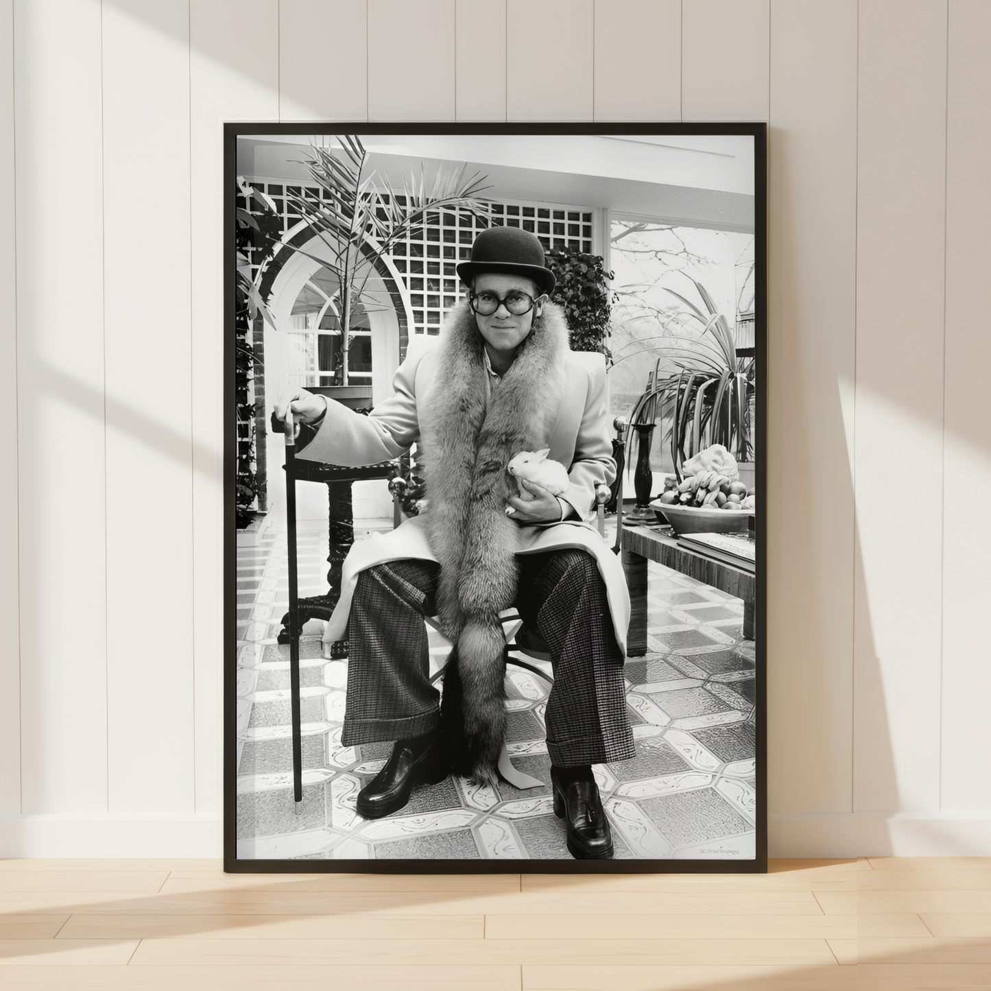 A framed black and white art print leaning against a white paneled wall on a light wood floor. The print shows a seated Elton John in a fur coat in an indoor architectural setting.