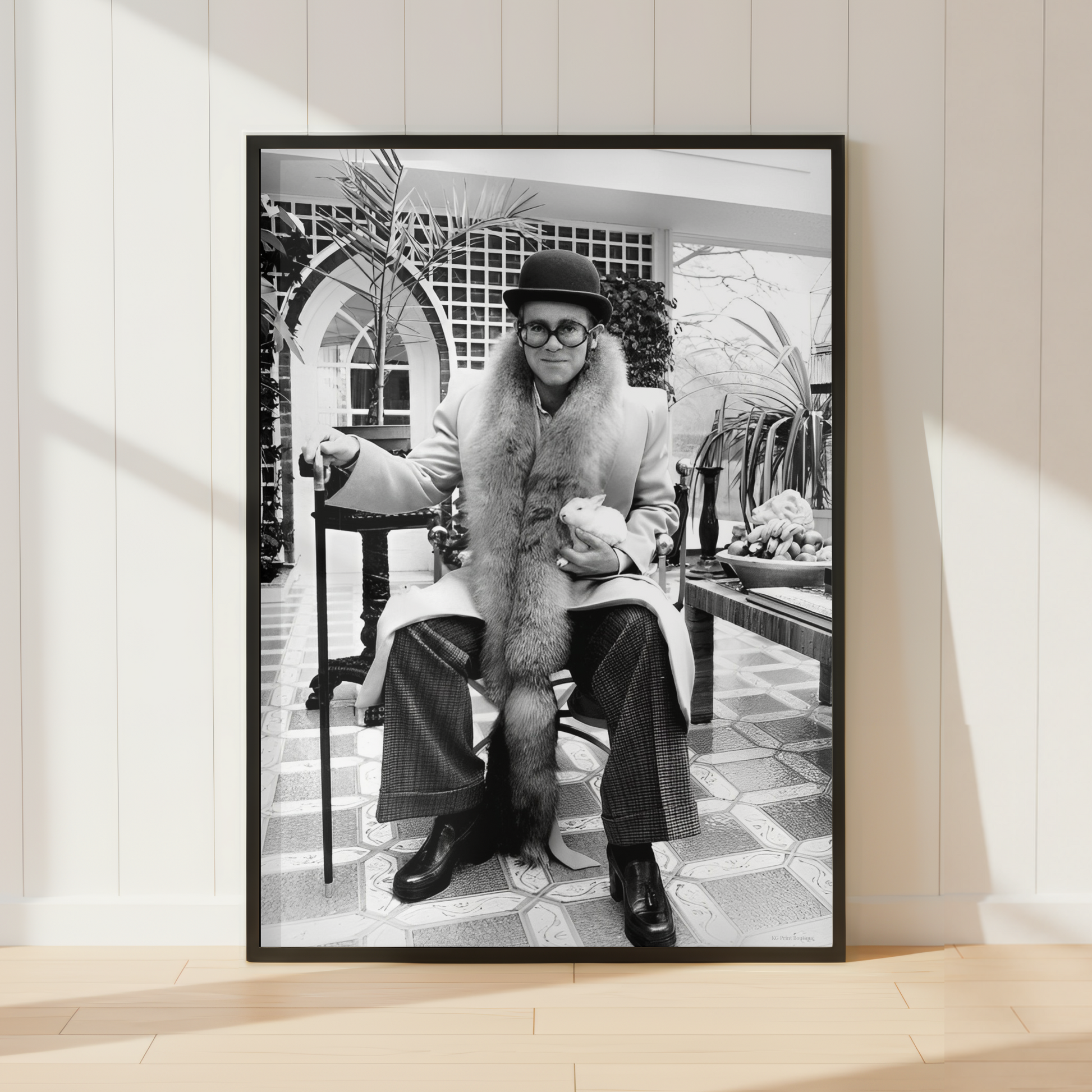 A framed black and white art print leaning against a white paneled wall on a light wood floor. The print shows a seated Elton John in a fur coat in an indoor architectural setting.