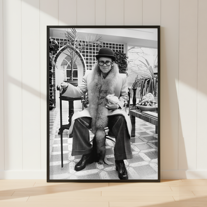 A framed black and white art print leaning against a white paneled wall on a light wood floor. The print shows a seated Elton John in a fur coat in an indoor architectural setting.