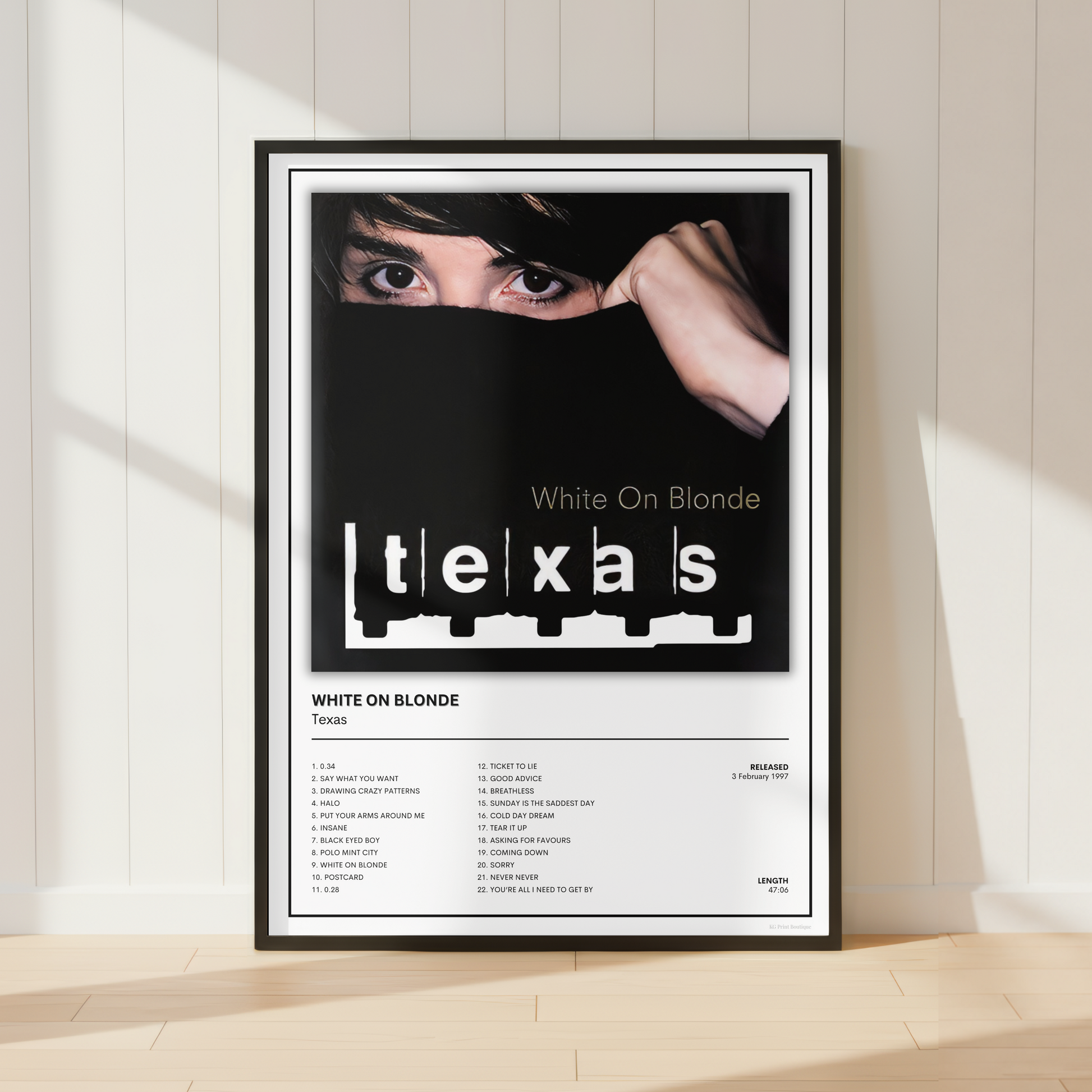A framed music poster for 'White On Blonde' by Texas stands against a white paneled wall on a light wood floor, with soft sunlight casting shadows.