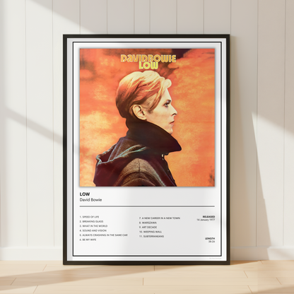 A framed David Bowie 'Low' music poster leaning against a white paneled wall on a light wood floor. The poster features a portrait of David Bowie in profile against a warm orange background, with the tracklist and album details printed below on a white section.