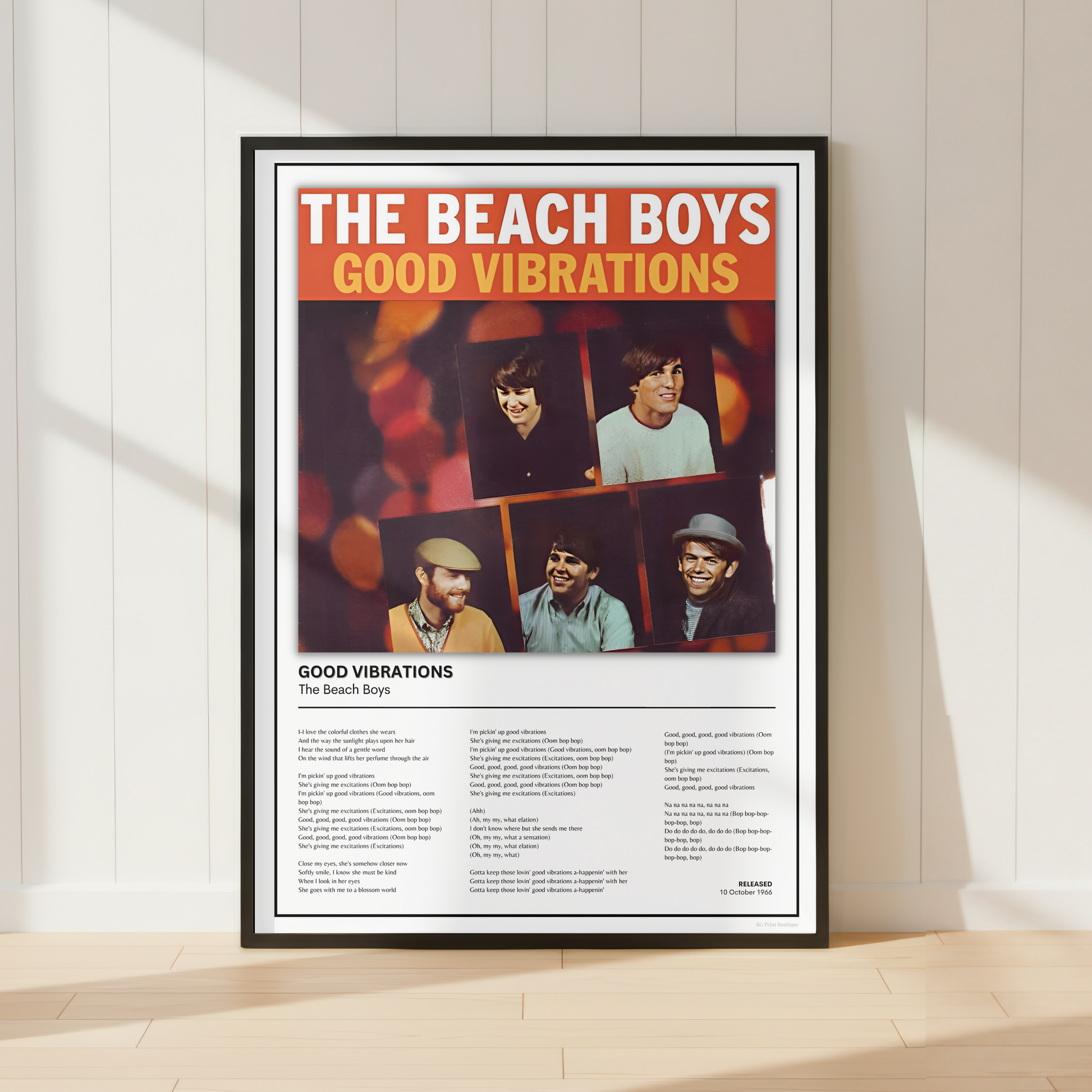 A framed Beach Boys 'Good Vibrations' poster leaning against a white paneled wall on a light wood floor, with soft sunlight casting shadows across the scene.