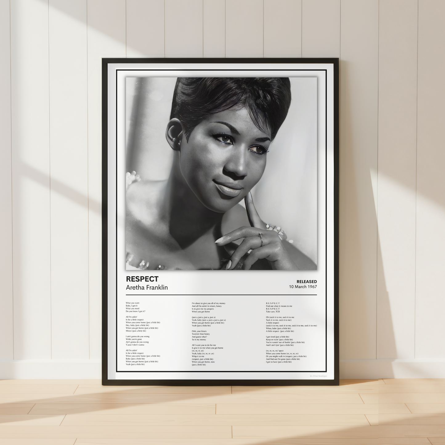 A framed black-and-white poster leaning against a cream paneled wall on a light wood floor. The poster features a portrait of Aretha Franklin with the title 'RESPECT' and song list text below.