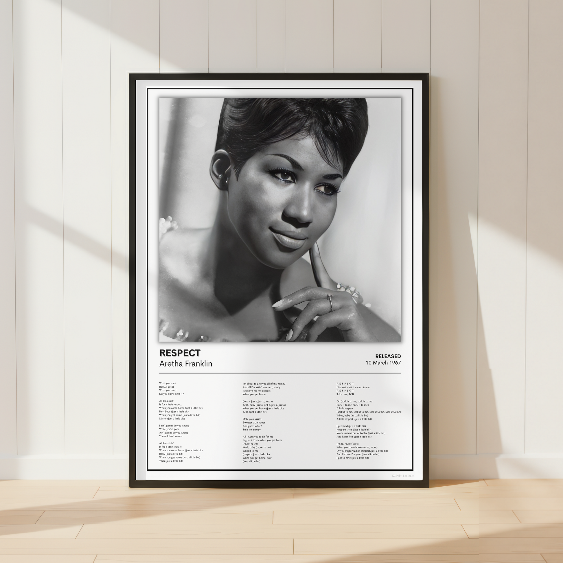 A framed black-and-white poster leaning against a cream paneled wall on a light wood floor. The poster features a portrait of Aretha Franklin with the title 'RESPECT' and song list text below.