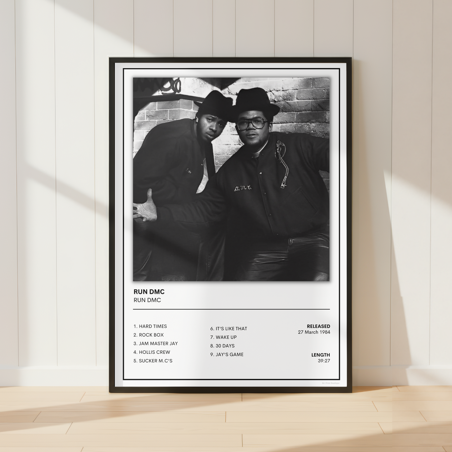 A framed black-and-white music poster for Run DMC leaning against a white paneled wall on a light wood floor, with soft sunlight casting shadows across the scene.