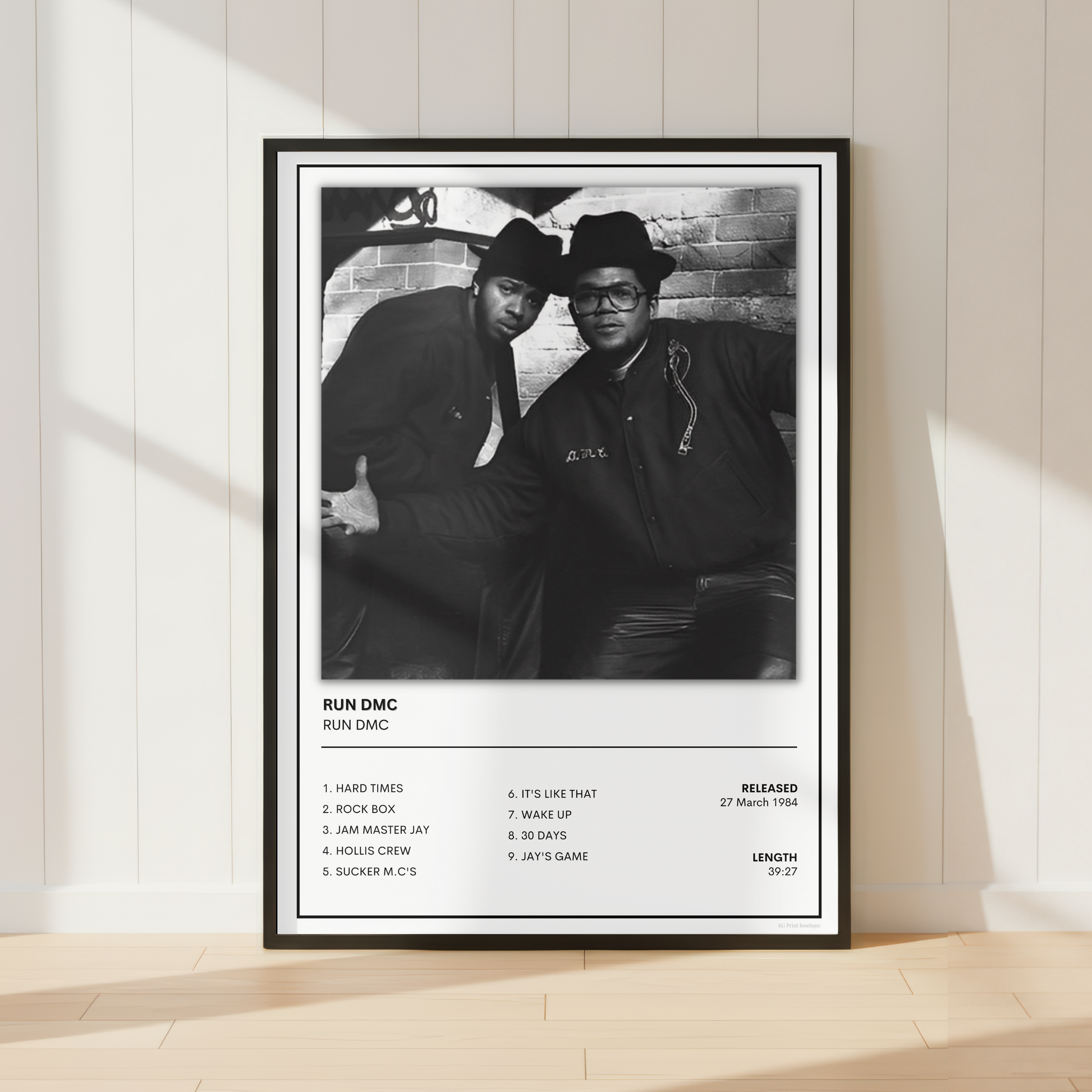 A framed black-and-white music poster for Run DMC leaning against a white paneled wall on a light wood floor, with soft sunlight casting shadows across the scene.