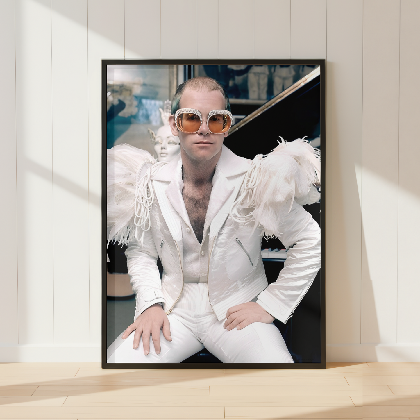 A framed vertical art print leaning against a white paneled wall on a light wood floor. The print shows Elton John wearing a white fringed jacket, white pants, and orange-tinted sunglasses.