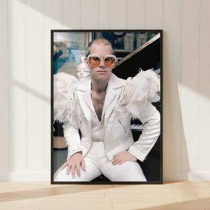 A framed vertical art print leaning against a white paneled wall on a light wood floor. The print shows Elton John wearing a white fringed jacket, white pants, and orange-tinted sunglasses.