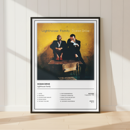 A framed Ocean Drive Lighthouse Family music poster is displayed leaning against a white paneled wall on a light wood floor, with soft sunlight casting shadows.