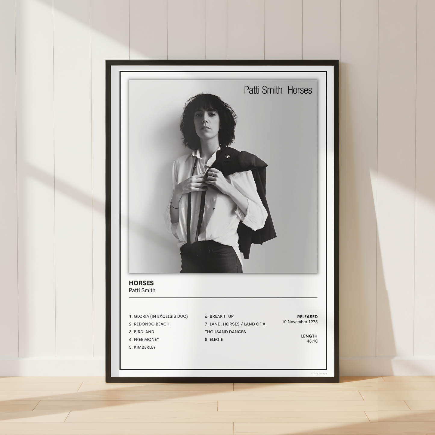 A framed black and white music poster of Patti Smith's album 'Horses' leaning against a cream-colored paneled wall on a light wood floor.