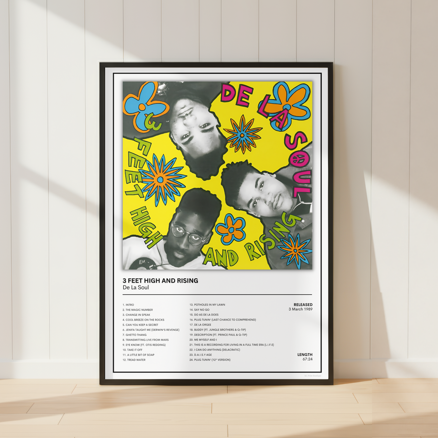 A framed music poster for De La Soul's '3 Feet High and Rising' leaning against a white paneled wall on a light wood floor.