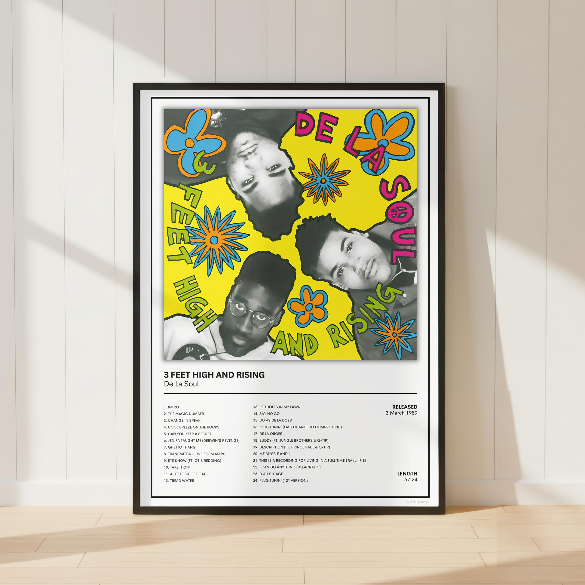 A framed music poster for De La Soul's '3 Feet High and Rising' leaning against a white paneled wall on a light wood floor.