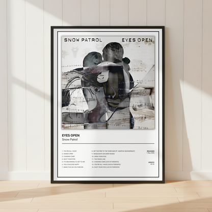 A framed Snow Patrol 'Eyes Open' music poster is displayed leaning against a white paneled wall on a light wood floor, with soft sunlight casting shadows.