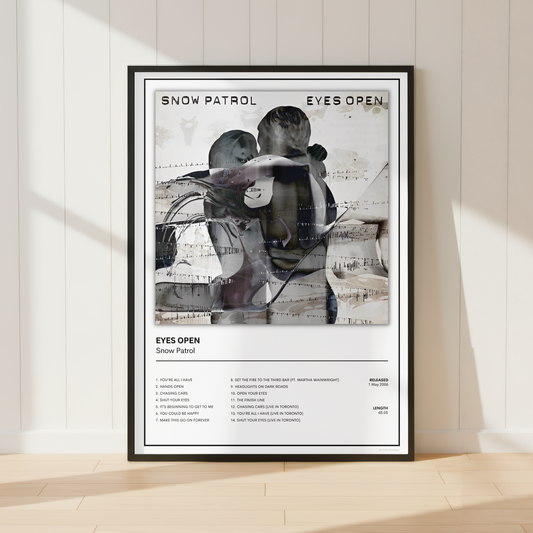 A framed Snow Patrol 'Eyes Open' music poster is displayed leaning against a white paneled wall on a light wood floor, with soft sunlight casting shadows.