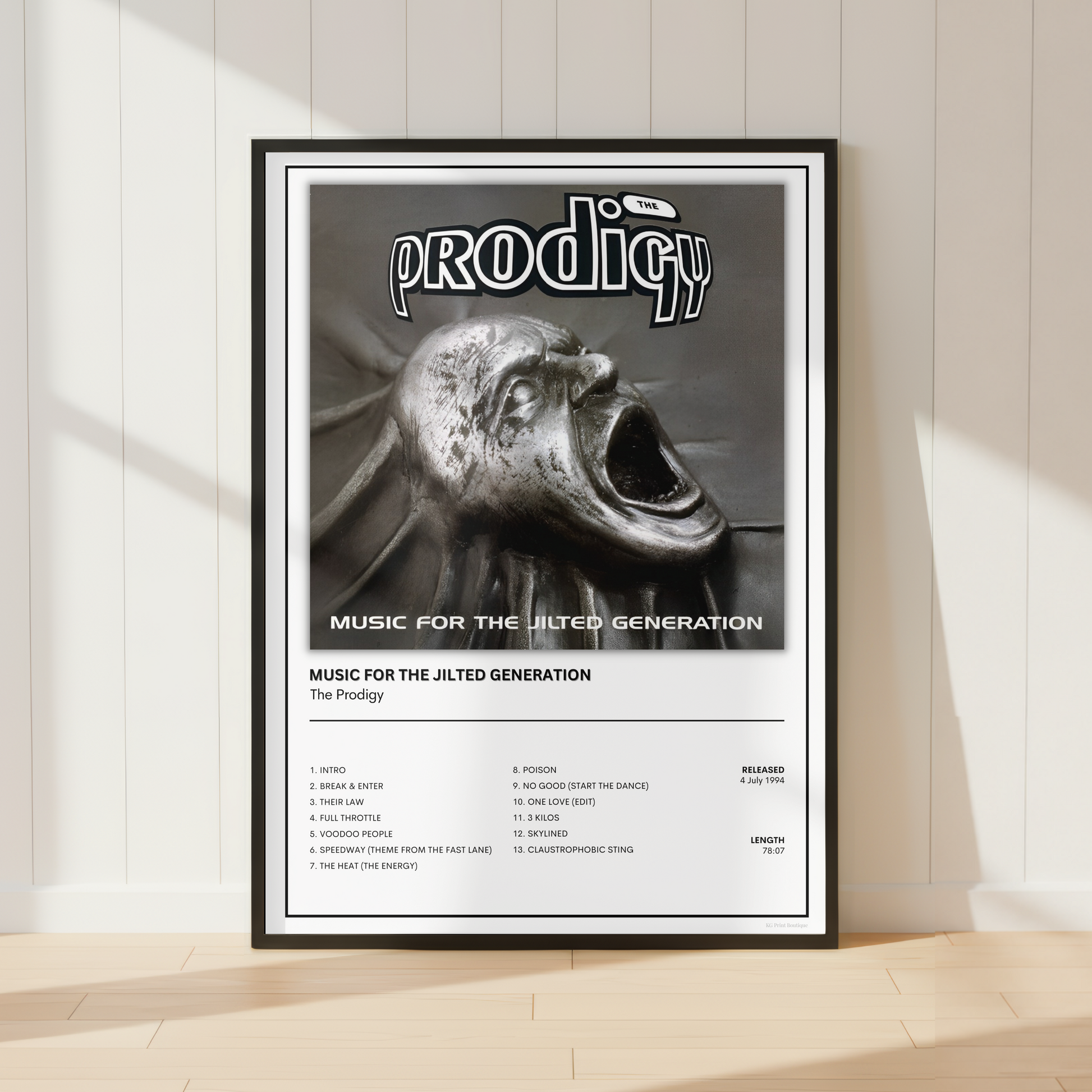 A framed music poster for the band Prodigy, featuring the album cover art for 'Music for the Jilted Generation' and a track list, leaning against a white paneled wall on a light wood floor.
