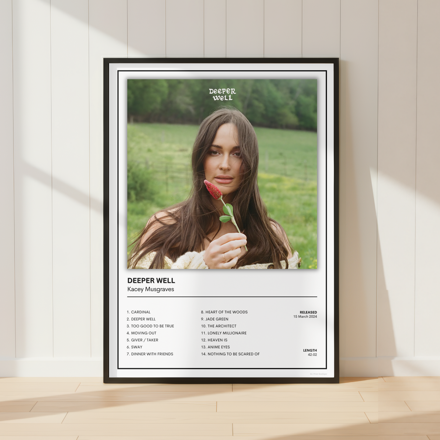 A framed music poster for Kacey Musgraves' album 'Deeper Well' leaning against a white paneled wall on a light wood floor. The poster features a portrait of Kacey Musgraves holding a red rose in a green field, with the album title and track list printed below.
