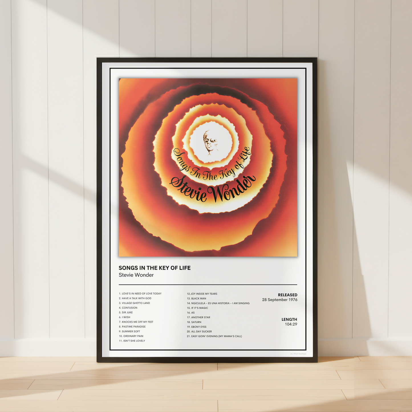 A framed music poster of Stevie Wonder's 'Songs in the Key of Life' leaning against a white paneled wall on a light wood floor, with soft window shadows.