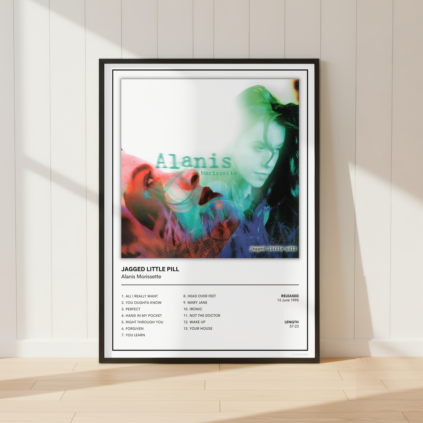 A framed music poster of Alanis Morissette's 'Jagged Little Pill' leaning against a white paneled wall on a light wood floor, with soft sunlight casting shadows.