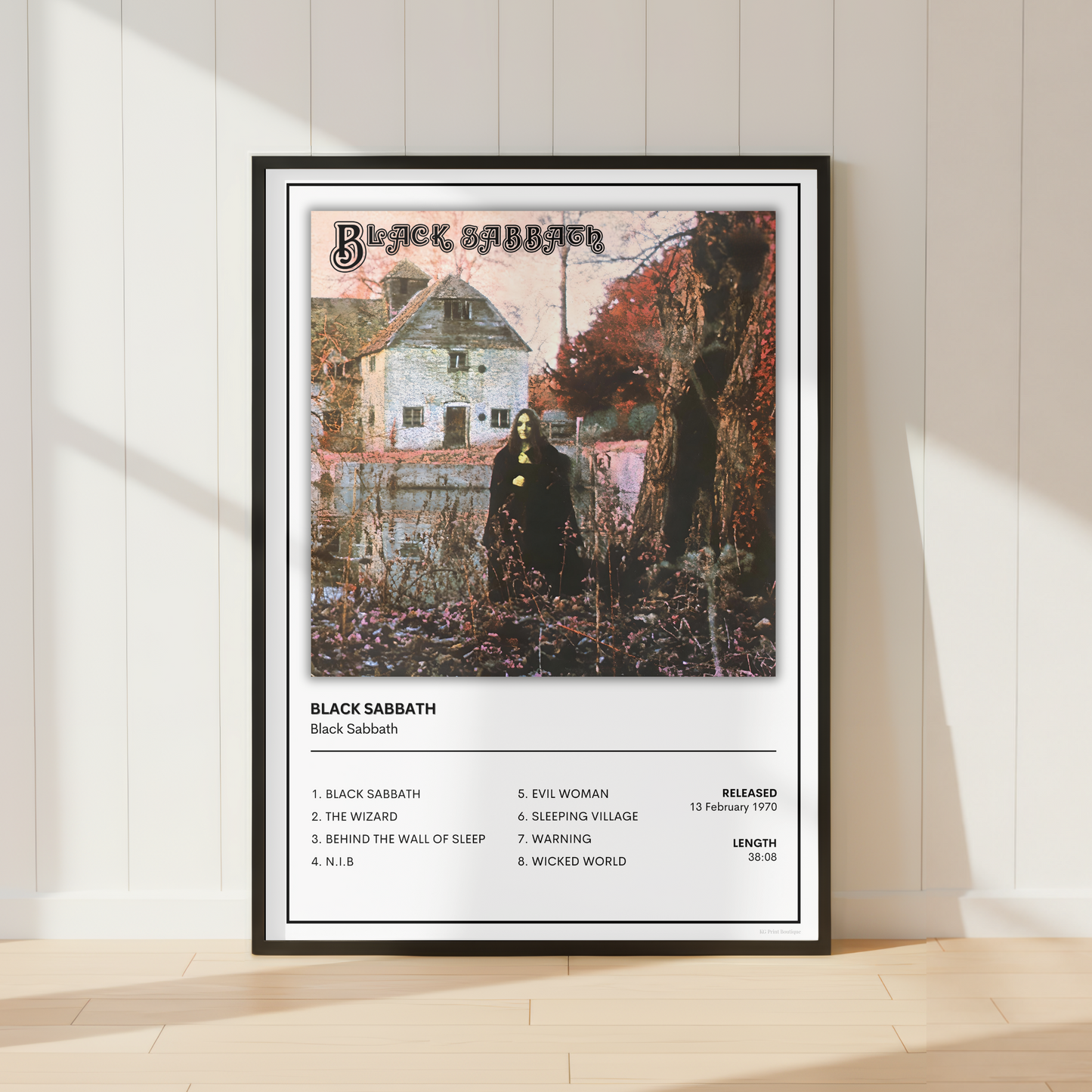 A framed Black Sabbath music poster leaning against a white paneled wall on a light wood floor, with soft sunlight casting shadows across the scene.