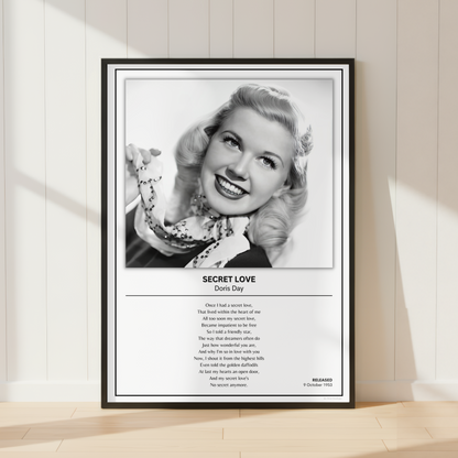 A framed black-and-white music poster of Doris Day's song 'Secret Love' leaning against a white paneled wall on a light wood floor, with soft sunlight casting shadows across the scene.