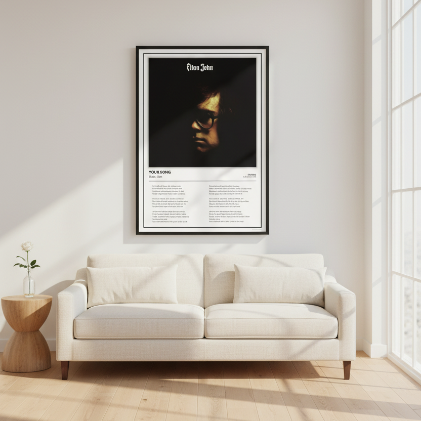 A framed Elton John song poster leaning against a white paneled wall on a light wood floor, with soft sunlight casting shadows across the scene.
