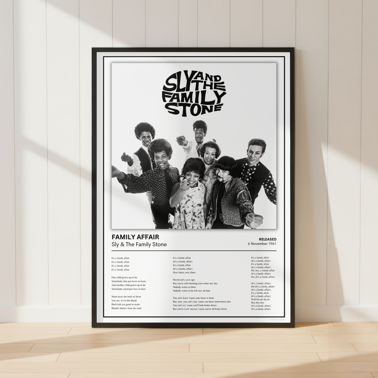 A framed black-and-white music poster for Sly and the Family Stone's 'Family Affair' leaning against a white paneled wall on a light wood floor, with soft window shadows cast across the scene.