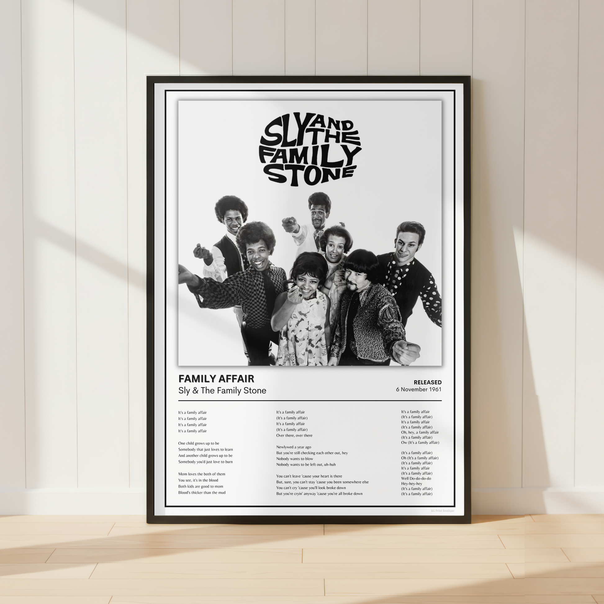 A framed black-and-white music poster for Sly and the Family Stone's 'Family Affair' leaning against a white paneled wall on a light wood floor, with soft window shadows cast across the scene.