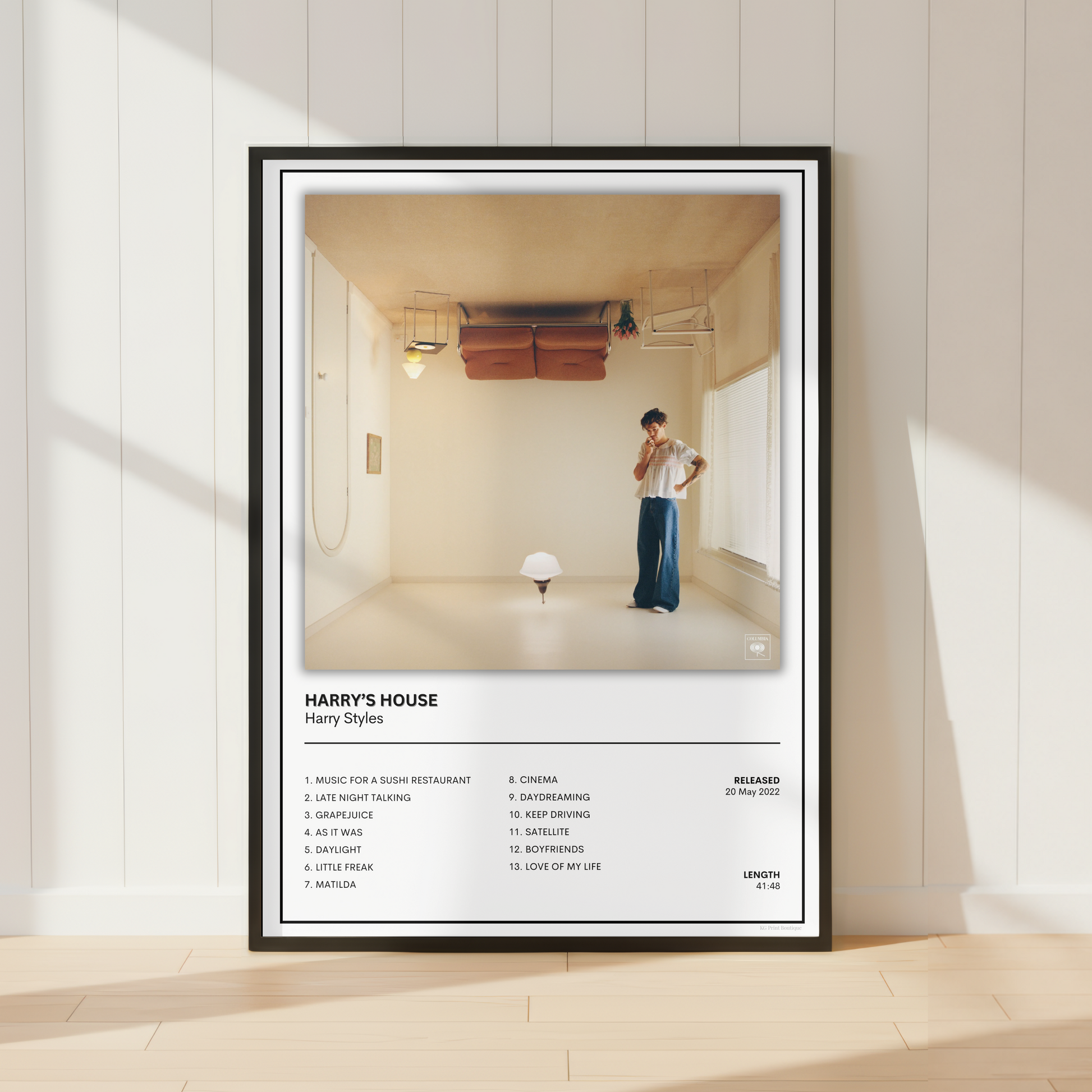 A framed music poster for Harry Styles' album 'Harry's House' leaning against a white paneled wall on a light wood floor. The poster features a black frame, white mat, and a photograph of a man in a minimalist room, with the tracklist printed below.