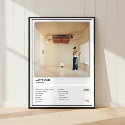 A framed music poster for Harry Styles' album 'Harry's House' leaning against a white paneled wall on a light wood floor. The poster features a black frame, white mat, and a photograph of a man in a minimalist room, with the tracklist printed below.